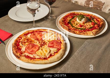 Schneiden Sie die Pizza mit natürlichen Tomatenscheiben und Ziegenkäse in der Nähe der Pizza mit Kirschtomaten und Parmesan, garniert mit Mozzarella-Käseball und Pesto-Sauce Stockfoto