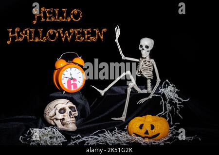 Ein Spielzeug Skelett kriecht aus dem Grab und winkt, Gruß, Kürbisse mit Grimassen, eine orangefarbene Uhr, ein Totenkopf auf schwarzem Hintergrund mit dem Text Er Stockfoto