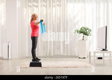 Ganzkörperaufnahme einer reifen Frau, die auf einem Aerobic-Stepper steht und zu Hause mit einem Gummiband vor dem fernseher trainiert Stockfoto