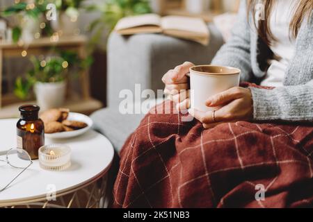 Kuschelige Frauenbeine im Winter-Strickpullover und kariertem Karomuschee trinken heißen Kakao oder Kaffee im Becher, sitzen zu Hause auf der Couch. Der Herbst vibes with Stockfoto