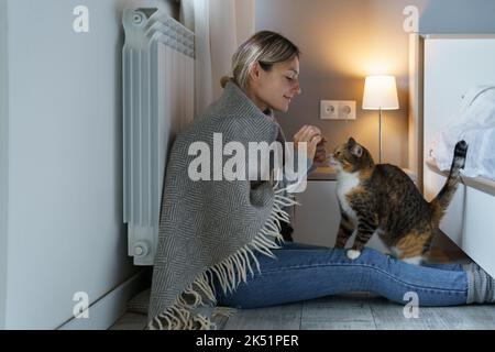Junge Frau mit warmer Decke bedeckt spielt mit Katze fütterndes Tier mit Essen auf dem Boden sitzen Stockfoto