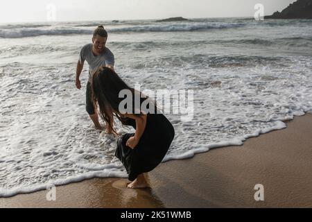 Bärtiger Mann, der lächelt, während er sich mit seiner Freundin im Kleid in der Nähe des Ozeans amüsieren kann, Stockbild Stockfoto