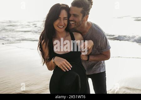 Fröhlicher Mann kitzelt freudige Freundin in Kleid in der Nähe des Ozeans in portugal,Stockbild Stockfoto