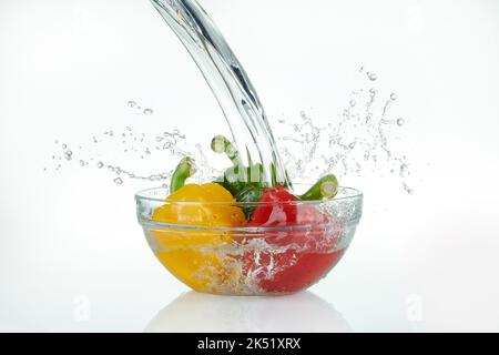 Wasserfall, der auf eine Glasschüssel mit bunten Paprika mit Spritzern auf weißem Hintergrund gießt. Stockfoto