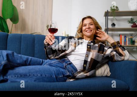 Eine fröhliche, angenehme Frau mittleren Alters mit einem Glas Wein in den Händen entspannt sich auf dem Sofa Stockfoto