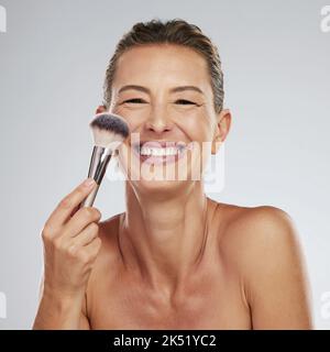 Schönheit, Make-up und Hautpflege Pinsel mit einer reifen Frau, die Kosmetik auf ihr Gesicht im Studio vor einem grauen Hintergrund. Kopfporträt einer Frau Stockfoto
