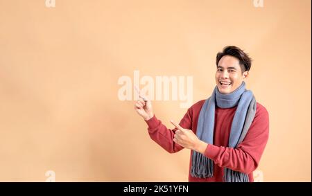 Positiver asiatischer Mann in rotem Pullover und Schal, der mit den Fingern auf einen leeren Bereich zeigt, der auf beigefarbenem Hintergrund isoliert ist. Fröhliche modische 20s Mann beiseite zeigen Geste - halbe Körperlänge Studio-Aufnahme. Stockfoto