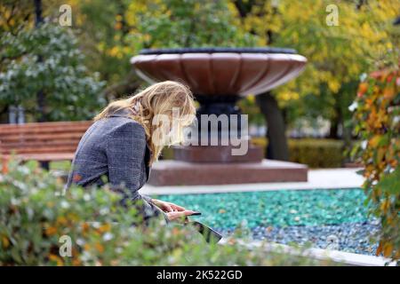 Frau mit blondem Haar im Mantel sitzt mit Smartphone auf einer Bank auf der Stadtstraße. Mit dem Handy im Herbstpark Stockfoto