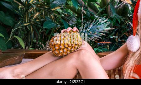 Kurzer Schuss von Frauenfüßen mit Ananas vor dem Hintergrund grüner tropischer Pflanzen. Frau im Hut des Weihnachtsmanns, die Ananas in den Händen hält. Konzept von Stockfoto