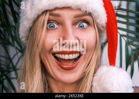 Lustige blonde Frau mit breitem Lächeln und Weihnachtsmütze, die mit überraschten Augen in die Kamera schaut. Mädchen feiert Weihnachten in den Tropen. Festliche Stimmung Stockfoto