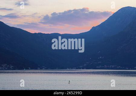 Entspannter junger Mann, der auf dem Boot rudert, mit Ruder, der den Sonnenuntergang auf dem Adriatischen Meer mit rosafarbenem Himmel und Bergen im Hintergrund betrachtet. Aktiver Lebensstil, Sommer. Stockfoto