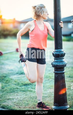 Echte junge reife kaukasische Frau in einem Sport tragen streching vor dem Laufen im Park an einem sonnigen Nachmittag. Fitnesskonzept. Stockfoto