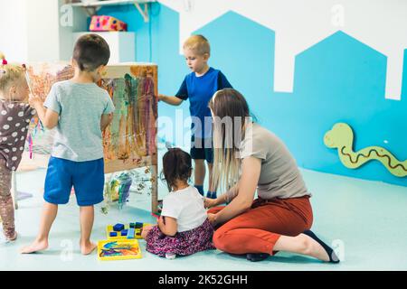 Vorschulkinder malen mit ihrer Lehrerin im Kindergarten auf das Zellophan, im Hintergrund schöne gemalte Wände. Hochwertige Fotos Stockfoto