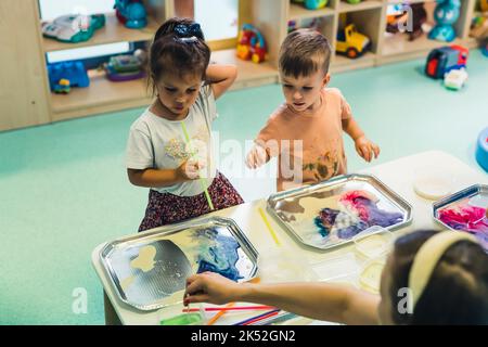 Vorschulkinder malen mit kleinen Tuben im Raum voller Spielzeug, Kindergarten mittellange Aufnahme. Hochwertige Fotos Stockfoto