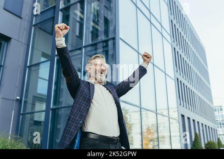 Erfolgreicher und glücklicher grauhaariger Chef vor dem Bürogebäude freut sich über Erfolg und Sieg, triumphiert mit Händen auf, Unternehmer-Unternehmer in Business-Anzug und Brille, reifen Investor Banker. Stockfoto