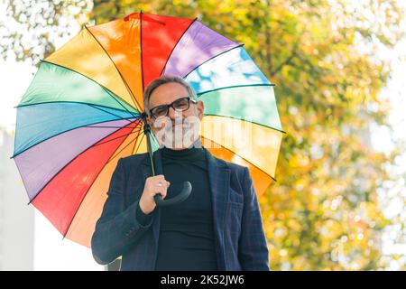 Schönes Outdoor-Porträt von positiven und lebensvollen älteren kaukasischen grauhaarigen Mann in Blazer und Rollkragen Blick auf die Kamera, während er seinen Kopf mit lebendigen Regenbogenschirm. Unscharfe Bäume mit gelben Blättern im Hintergrund. Hochwertige Fotos Stockfoto