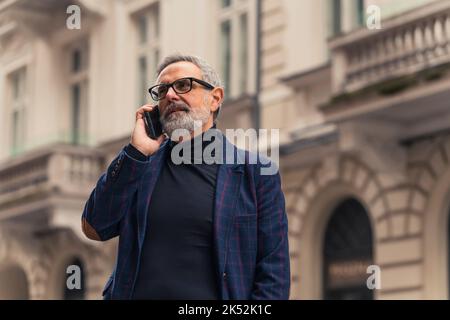 Mitten im Freien Aufnahme eines in Business-Freizeit gekleideten älteren kaukasischen Mannes mit grauem Gesichtshaar und schwarzer Brille, der mit seinem modernen Smartphone spricht. Elegantes Gebäude im Hintergrund verschwommen. Hochwertige Fotos Stockfoto