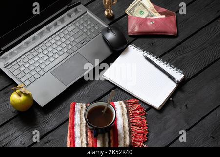 Schwarzer Laptop-Notizblock-Becher mit Kaffeegeld liegt auf dem Arbeitstisch aus schwarzem Holz, Laptop während der Arbeit auf dem Arbeitstisch, kreativer Prozess, freiberuflich Stockfoto