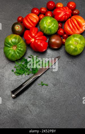 Verschiedene Sorten von roten und grünen Tomaten. Gehackte Petersilie und Messer. Speicherplatz kopieren. Flach liegend. Schwarzer Hintergrund. Stockfoto