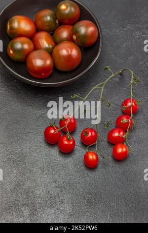 Zweig aus Kirschtomaten, rote Tomaten in schwarzer Schüssel. Speicherplatz kopieren. Flach liegend. Schwarzer Hintergrund. Stockfoto