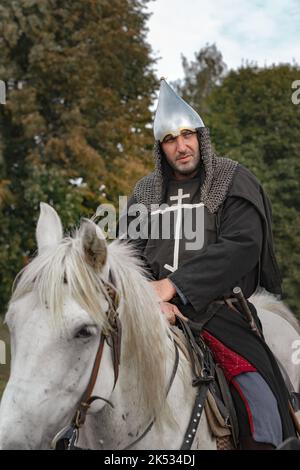Reiter in Kettenpanzerung, Helm und christliches Kreuz auf einer Robe auf weißem Pferd an bewölktem Tag Stockfoto