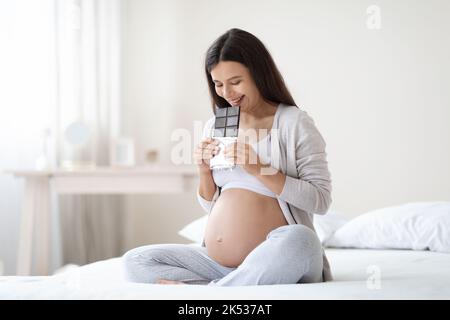 Glückliche schöne Schwangere, die auf dem Bett sitzt und Schokolade isst Stockfoto