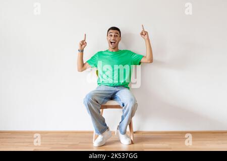 Aufgeregter Mann aus dem Nahen Osten, der mit zwei Fingern nach oben zeigt, auf einem Stuhl auf einer weißen Studiowand sitzend, für dein Werbedesign Stockfoto