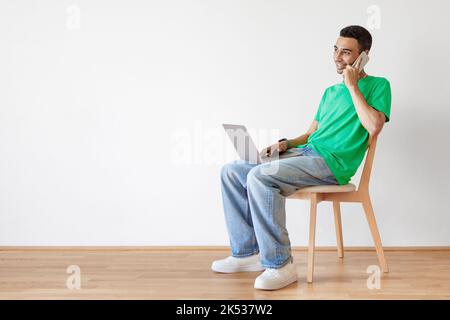 Positiver arabischer Mann, der auf einem Stuhl mit Computer auf dem Schoß sitzt, mit dem Arbeitgeber telefoniert und Platz kopiert Stockfoto