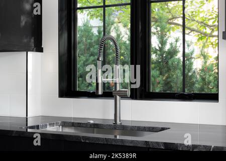 Ein Küchendetail mit schwarzen Schränken und Marmorarbeitsplättchen, weißen U-Bahnfliesen und Edelstahlhahn und Waschbecken vor einem Fenster. Stockfoto