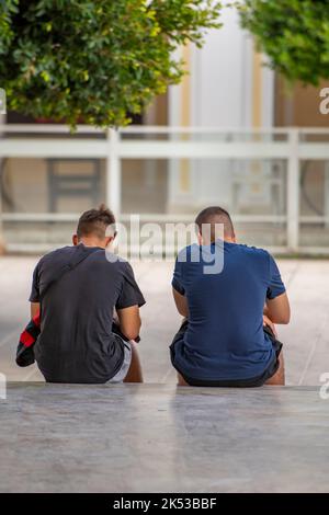 Zwei junge Jungs, die mit Smartphones an der Wand saßen, zwei Freunde, die mit Smartphones spielten, junge Männer, die Technologie nutzten, zwei Jungen, die in Telefone vertieft waren. Stockfoto