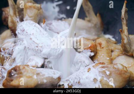 Der Koch bereitet Huhn - Beine und Flügel im Freien zu. Milch - Rahm wird mit gebratenem Huhn in einen Kessel gegossen. Nahaufnahme. Stockfoto