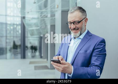 Glücklicher, gutaussehender grauhaariger Mann, Geschäftsmann in Anzug und Brille. Hält das Telefon in den Händen, erhält gute Nachrichten, liest die Nachricht. Es liegt an der Straße in der Nähe eines modernen Gebäudes Stockfoto