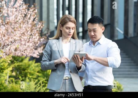 Interrassische Partnerschaft. Junge Geschäftsfrau und junger asiatischer Geschäftsmann, der in der Nähe des Bürozentrums steht, spricht, am Tablet arbeitet, über Geschäfte diskutiert, Zusammenarbeit, Vereinbarung. Stockfoto