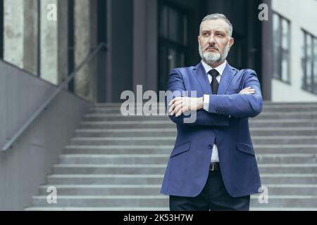 Porträt eines älteren, grauhaarigen Leiters und ceo auf der Treppe des Bürozentrums. Er steht ernst in einem Anzug, die Arme sind vorne gekreuzt und blicken zur Seite. Stockfoto