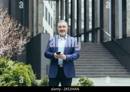 Ein gutaussehender grauhaariger Mann in einem Anzug geht zu einer Geschäftsbesprechung, nutzt das Telefon auf der Straße, hält es in den Händen, wählt. Er schaut lächelnd auf die Kamera. Stockfoto