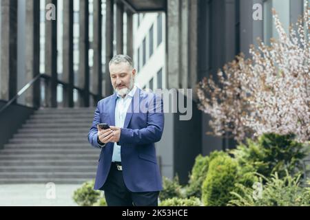 Ein älterer grauhaariger Mann, ein Geschäftsmann in einem Anzug, steht in der Nähe des modernen Bürozentrums und ruft vom Telefon aus ein Taxi, tippt die Nummer ein, wartet, ist eilig, sich zu treffen. Stockfoto