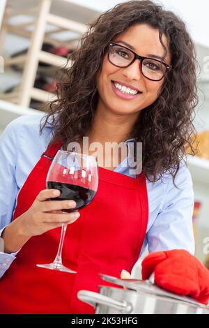 Schöne glückliche hispanische Latina junge Frau oder Mädchen mit perfekten Zähnen trägt eine rote Schürze trinken ein Glas Rotwein und Kochen in ihrer Küche an Stockfoto