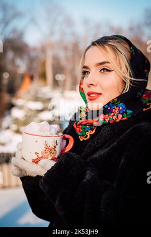 Ein Mädchen in einem ukrainischen Nationalschal hält im Winter eine Tasse Marschmallchen Stockfoto