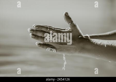 Die Priesterin gießt sauberes Wasser aus. Handflächen mit Zigeuner-Boho-Ringen mit klarem Aqua. Meditation, Beten, Dankbarkeit, Reinheit Hintergrund. Schwarz und Stockfoto
