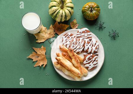 Teller mit Halloween-Keksen, Glas Milch, Blättern und Kürbissen auf grünem Hintergrund Stockfoto