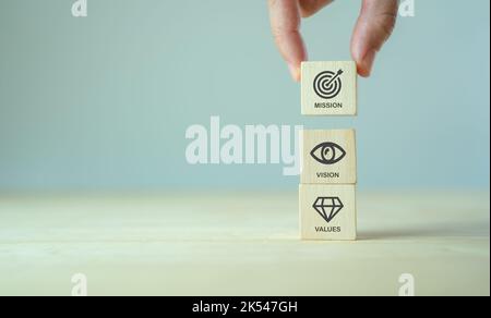 Mission, Vision und Werte des Unternehmens. Zweck Geschäftskonzept. Hand hält Holzwürfel mit Mission Vision und Werte Symbole auf grauem Hintergrund. Mod Stockfoto