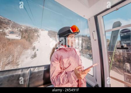 Skiurlaub - Frau Skifahrerin mit Telefon-App in Gondelbahn. Lächelndes Mädchen mit dem mobilen Smartphone, das Skibekleidung, Helm und Brille trägt. Ski Stockfoto