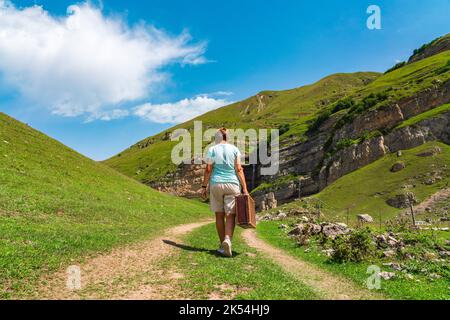 Ein Mädchen mit einem Koffer geht auf einer unbefestigten Straße in den Bergen. Interessante Reise der einsamen Frau Stockfoto