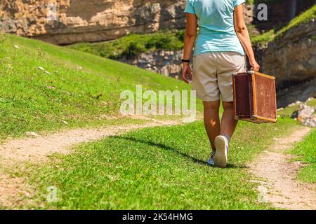 Ein Mädchen mit einem Koffer geht auf einer unbefestigten Straße in den Bergen. Interessante Reise der einsamen Frau Stockfoto