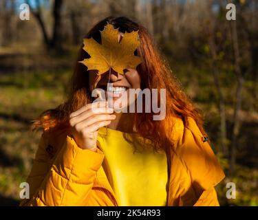 Rothaarige kaukasische Frau, die ein gefallenes Ahornblatt hält. Herbstspaziergang. Stockfoto