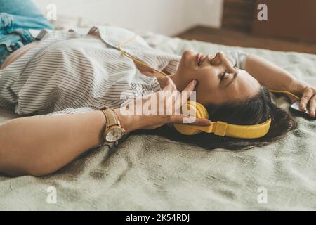 Eine schöne Frau mit dunklem Haar liegt zu Hause in Kopfhörern auf dem Bett. Stockfoto