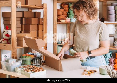 Lächelnd konzentrierte lockige Frau Verpackung und Verpackung Versand mit Silikon Babyschale in Karton zur Lieferung. Holzrassel Spielzeug, trinket Stockfoto