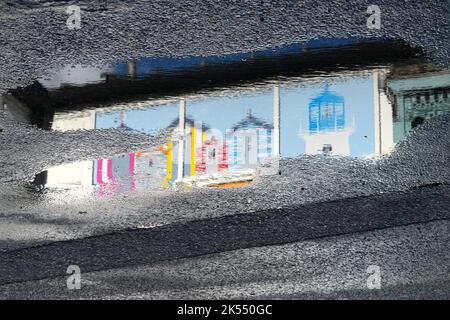 Wandmalereien am Meer von Strandhütten und Leuchtturm spiegeln sich auf der nassen Straße am Mumbles Pier, nachdem der Regen aufgeräumt hat. Stockfoto