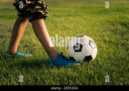 Klassischer schwarz-weißer Fußballball und Beine eines Fußballers auf dem grünen Gras des Feldes. Fußballspiel, Training, Hobby-Konzept. Mit Kopie-SPAC Stockfoto