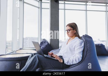 Fröhliche blonde Frau im weißen Hemd, die mit einem Laptop-Computer einen Veröffentlichungstext schreibt, ein fröhliches Mädchen mit Brille, das aus der Ferne arbeitet, teilt einen positiven Betrug Stockfoto